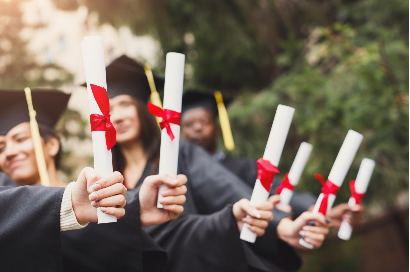 Zdjęcie przedstawia grupę absolwentów ubranych w togi i birety, trzymających w wyciągniętych rękach zrolowane dyplomy przewiązane czerwonymi wstążkami.