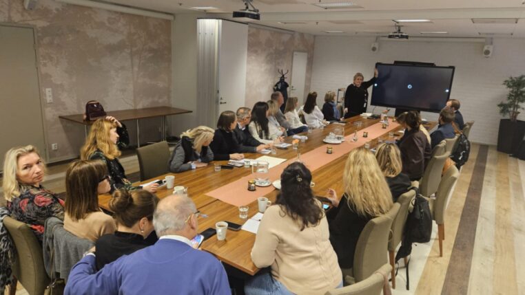 Grupa osóbsiedząca przy stole, słuchająca wykładowczyni, która stoi przy ekranie telewizyjnym.