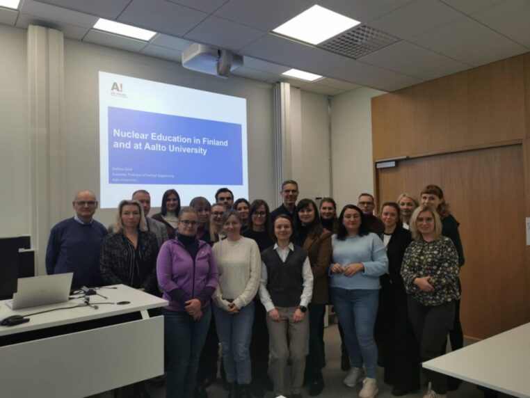 Grupa stojących na tle prezentacji dotyczącej edukacji o energetyce jądrowej, uczestników wizyty wraz panią profesor z Uniwersytetu Aalto.