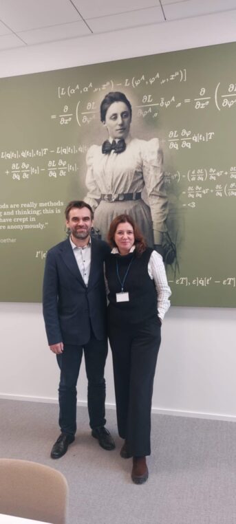 Dwie osoby - kobieta i mężczyzna stojące na tle wizerunku Emmy Noether - niemieckiej matematyczki i fizyczki