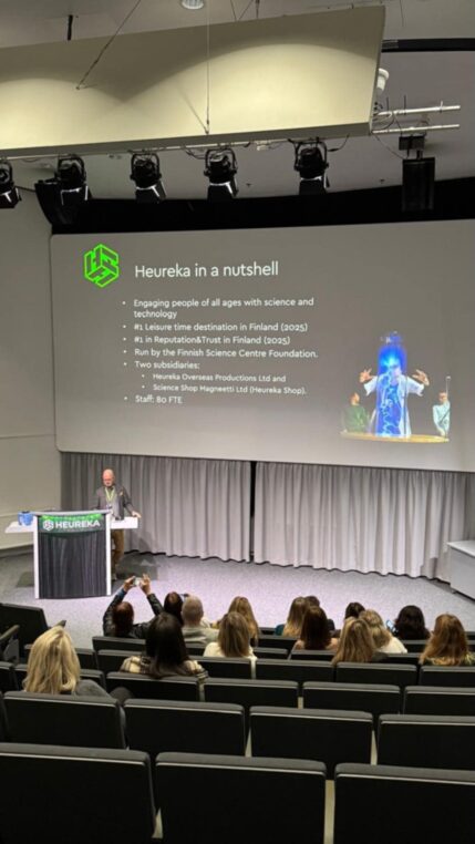 Uczestnicy siedzą i oglądają wyświetlaną prezentację, która przedstawia stojący przy mównicy wykładowa.