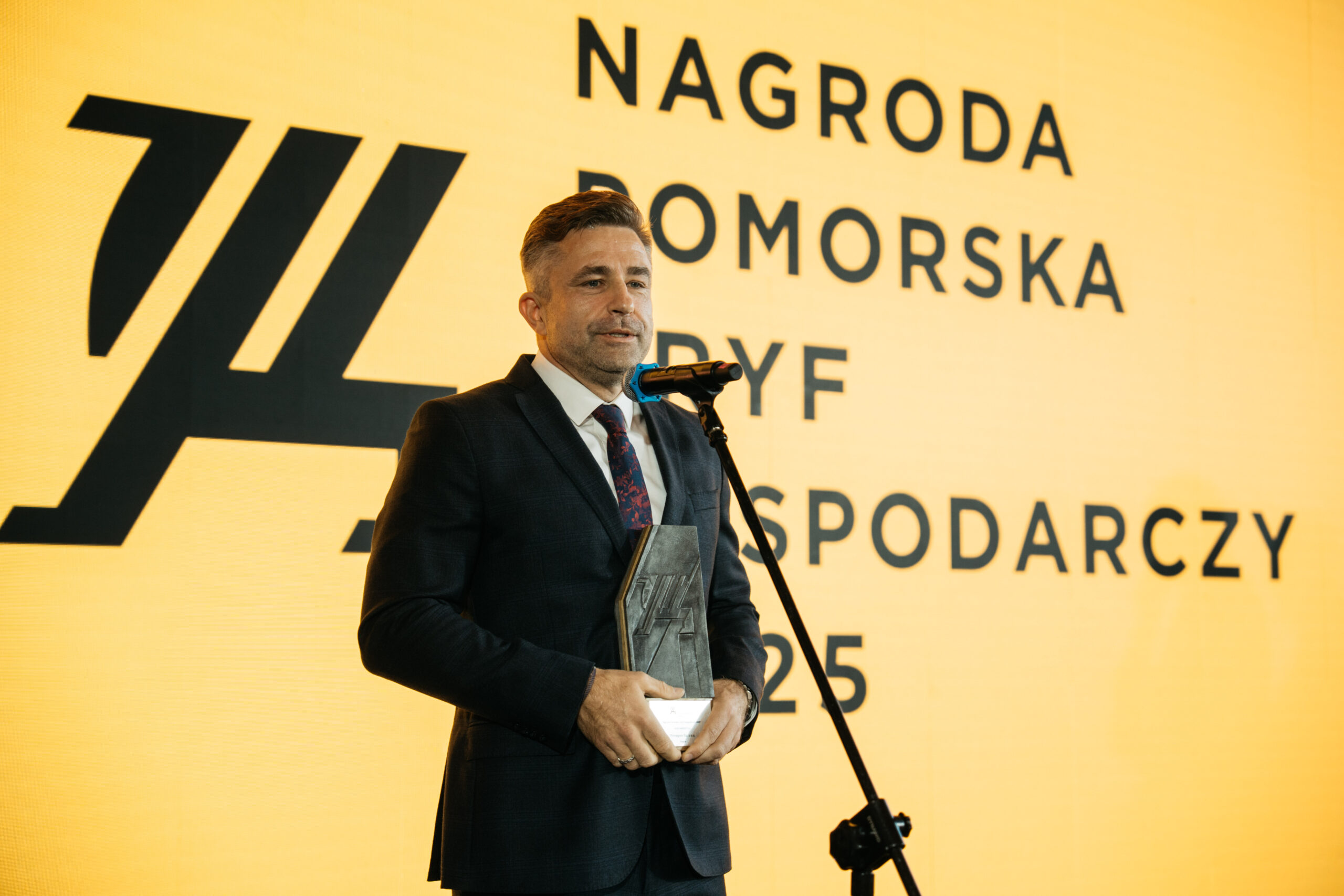 Fotografia przedstawiająca Tomasza Sznajdera, prezesa Primagran, mówiącego do mikrofonu, na tle ekranu wyświetlającego logotyp Gryfa Gospodarczego