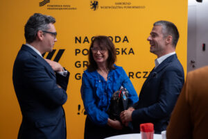 Zdjęcie trzech osób na tle ścianki konkursu gryf gospodarczy. od lwesj, mężczyzna (profilem, kobieta (frontem), mężczyzna (profilem)